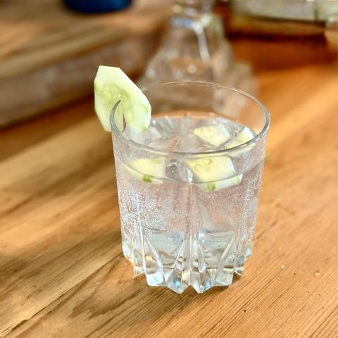 A gin and tonic garnished with cucumber sitting on a wooden table.